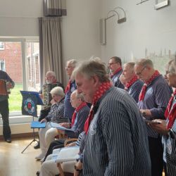 Eine Gruppe älterer Männer singt fröhlich in einem Raum, während ein Mann im Hintergrund das Akkordeon spielt.