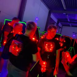 Menschen spielen Laser-Tag in einer dunklen Arena mit leuchtenden Westen und roten Lichtern zwischen Hindernissen.