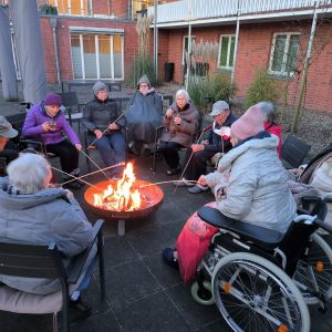 Ältere Menschen sitzen gemütlich im Innenhof um ein Lagerfeuer und grillen gemeinsam Marshmallows.