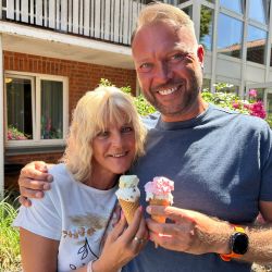 Ein lächelndes Paar genießt an einem sonnigen Tag zwei Eiswaffeln vor einem Gebäude mit Blumen im Hintergrund.