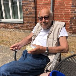 Ein älterer Mann sitzt entspannt vor einer Backsteinwand und genießt ein Dessert im Freien.