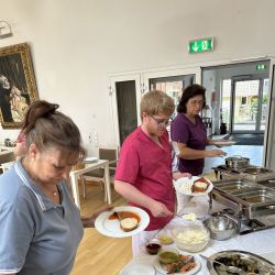 Drei Personen servieren Essen an einem Buffet mit vielfältigen Speisen in einem hell erleuchteten Raum.