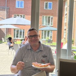 Ein Mann sitzt drinnen und genießt ein Stück Pizza, während durch die Fenster ein sonniger Innenhof sichtbar ist.