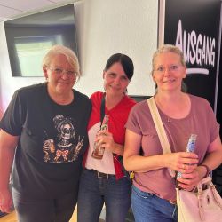 Drei Frauen posieren fröhlich mit Getränken vor einem Schild mit "Ausgang" und genießen den Moment.