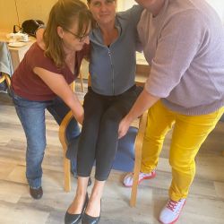 Zwei Personen helfen einer lächelnden Frau, die auf einem Stuhl sitzt, während alle gemeinsam in die Kamera schauen.