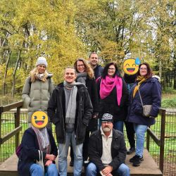 Eine Gruppe von Menschen posiert lächelnd auf einer Holzplattform im herbstlichen Wald vor bunt gefärbten Bäumen.