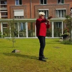 Ein Mann in rotem Sakko singt im Garten vor einem Backsteingebäude, während Menschen ihm im Sonnenschein zuhören.
