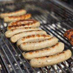 Mehrere Bratwürste mit goldbrauner Kruste brutzeln auf einem Grill in der Sommersonne.