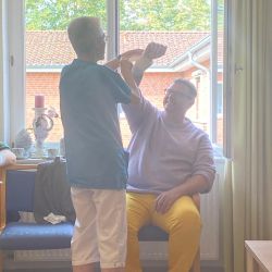 Eine Person hebt den Arm einer sitzenden Person in einem sonnendurchfluteten Raum mit Blick auf ein rotes Ziegelhaus an.