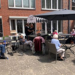 Senioren genießen Eis und Unterhaltung im Freien unter einem großen Sonnenschirm vor einem Backsteingebäude.