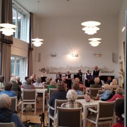 Senioren genießen ein Konzert im Gemeinschaftsraum mit großen Fenstern und einer beeindruckenden Wandmalerei im Hintergrund.