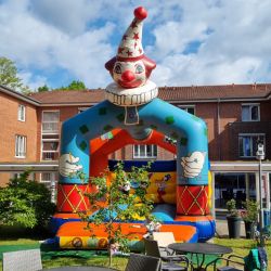 Eine bunte Hüpfburg in Clownform steht in einem sonnigen Garten mit leeren Tischen und Stühlen im Vordergrund.