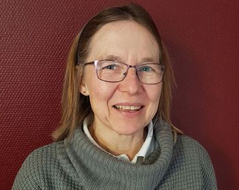 Eine lächelnde Frau im grauen Pullover steht vor einer dunkelroten Wand und strahlt Wärme und Freude aus.