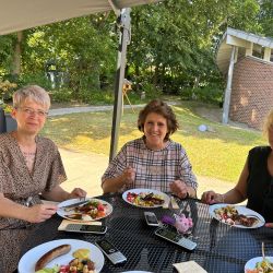 Drei Frauen sitzen unter einem Sonnenschirm und genießen ein Sommeressen mit Salat und Würstchen im Freien.