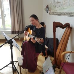 Eine Frau spielt Gitarre und singt in einem gemütlichen Raum mit Harfe und Notenständer vor sich.