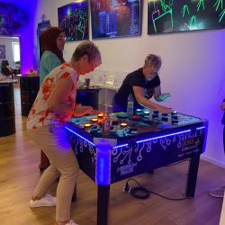 Drei Personen spielen gespannt ein interaktives Spiel auf einem beleuchteten Tisch in einer modernen Spielhalle.