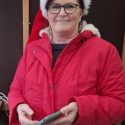 Eine Frau in rotem Mantel und Weihnachtsmütze lächelt fröhlich, während sie ein Smartphone in einem gemütlichen Raum hält.