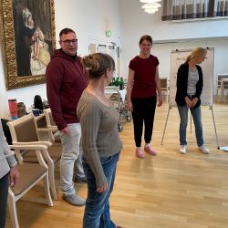 Eine Gruppe von Menschen steht in einem Raum und scheint an einem Workshop teilzunehmen, während sie miteinander kommunizieren.