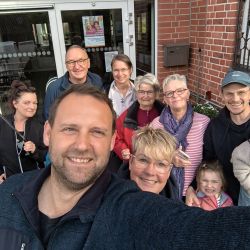 Eine Gruppe von Menschen macht ein fröhliches Selfie vor einer Backsteinfassade eines Gebäudes an einem sonnigen Tag.
