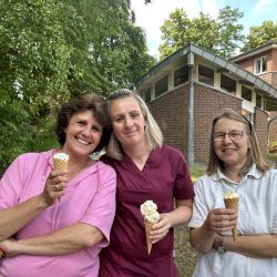 Drei Frauen genießen bei sonnigem Wetter Eiscreme vor einem Backsteingebäude mit üppigem Grün im Hintergrund.
