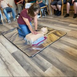 Eine Frau demonstriert Erste-Hilfe-Maßnahmen an einer Puppe, während mehrere Personen aufmerksam im Hintergrund zuschauen.