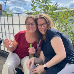 Zwei Frauen sitzen fröhlich draußen, trinken Getränke und genießen das sonnige Wetter vor einer grünen Kulisse.