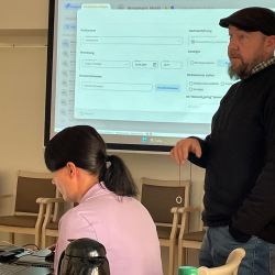 Ein Mann präsentiert Informationen auf einem Bildschirm, während eine Frau am Laptop arbeitet, bei einem Workshop.