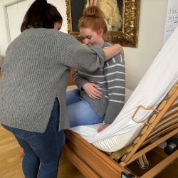 Zwei Frauen interagieren freundlich, während eine auf einem verstellbaren Bett in einem hellen Raum sitzt.