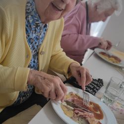 Eine ältere Frau in gelber Jacke genießt lächelnd ihr Mittagessen, während eine andere Person im Hintergrund sitzt.