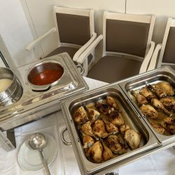 Auf einem weißen Tischtuch steht ein Buffet mit Brathähnchen und Saucen, flankiert von drei braunen Stühlen.