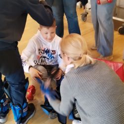 Eine Frau hilft einem Jungen, Skischuhe anzuziehen, während umstehende Personen neugierig zuschauen.