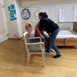 Eine Frau in Jeans hilft einer sitzenden Person, von einem Stuhl neben einem Pflegebett in einem hellen Raum aufzustehen.