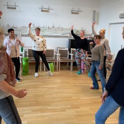 Eine Gruppe von Menschen tanzt fröhlich in einem Raum mit Holzboden und hebt die Arme in die Luft.