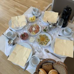 Ein reich gedeckter Frühstückstisch mit Brötchen, Lachs, Ananas und Aufschnitt steht auf einem hellen Holzboden.