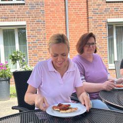 Zwei Frauen sitzen draußen an einem Tisch, während eine lächelnd ein Stück Pizza auf einem Teller präsentiert.