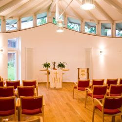 Ein heller Kapellenraum mit hölzernem Boden und roten Stühlen ist für einen bevorstehenden Gottesdienst vorbereitet.