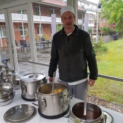 Ein Koch in Arbeitskleidung bereitet in einem verglasten Raum eine Mahlzeit an großen Suppentöpfen zu.