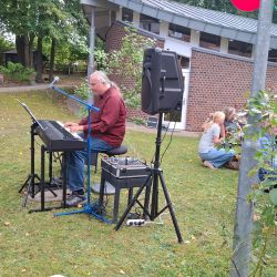 Ein Mann spielt im Freien Keyboard, während Menschen entspannt auf dem Rasen sitzen und der Musik lauschen.