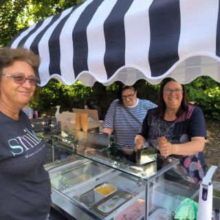 Drei Frauen stehen fröhlich an einem Eisstand mit gestreiftem Sonnendach und genießen einen sonnigen Tag im Freien.