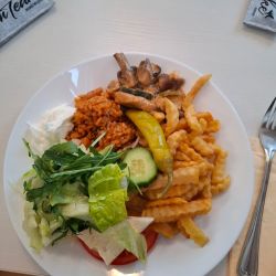 Ein Teller mit Pommes, Reis, Salat, Pilzen und einer Peperoni liegt auf einem Tisch mit Besteck und Servietten.