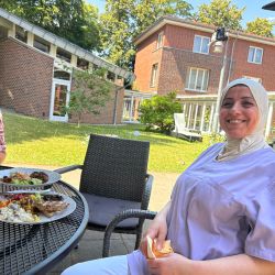 Eine lächelnde Frau in Arbeitskleidung genießt ihr Essen im Freien vor einem Backsteingebäude.