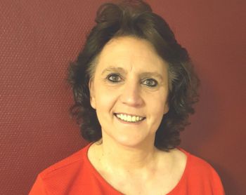 Eine Frau mit braunen Haaren lächelt herzlich vor einem kräftig roten Hintergrund in die Kamera.