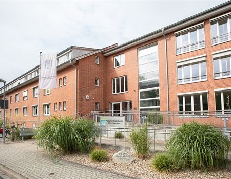 Ein großes Backsteingebäude mit vielen Fenstern steht unter einem bewölkten Himmel, umgeben von grünen Pflanzen.