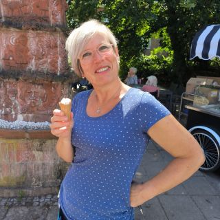 Eine Frau in einem blauen Punkteshirt genießt ein Eis vor einem kunstvoll verzierten Backsteinturm an einem sonnigen Tag.