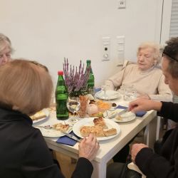 Eine Gruppe älterer Menschen genießt Pizza an einem festlich dekorierten Tisch mit Getränken und Pflanzen.