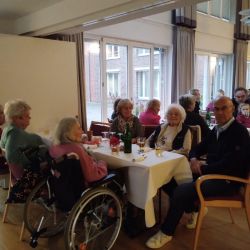 Ältere Menschen und Begleiter genießen geselliges Beisammensein an einem festlich gedeckten Tisch in einem hellen Raum.