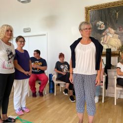 Eine Gruppe von Erwachsenen steht und sitzt entspannt auf einem Holzboden vor einem großen, gerahmten Gemälde an der Wand.