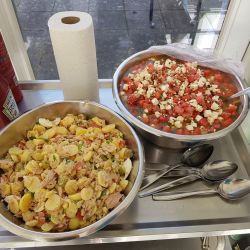 Zwei Schüsseln mit Kartoffelsalat und Tomaten-Feta-Salat stehen auf einem Tisch, umgeben von Ketchup- und Senf-Flaschen.
