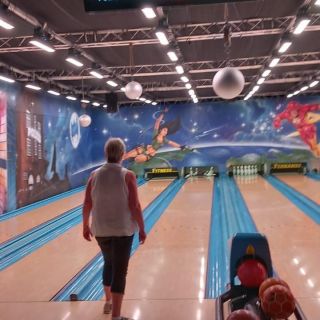 Eine Person geht auf der Bowlingbahn, während ein Bildschirm die aktuellen Spielstände von Chris, Michel und anderen anzeigt.