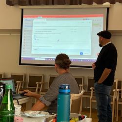 Ein Mann hält eine Präsentation vor einer Projektionsleinwand, während ein anderer am Laptop arbeitet.